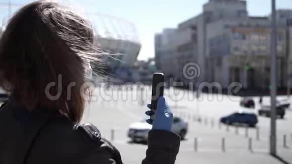 戴着外科口罩和手套的年轻女性在冠状病毒流行期间在户外智能手机上录制视频视频的预览图