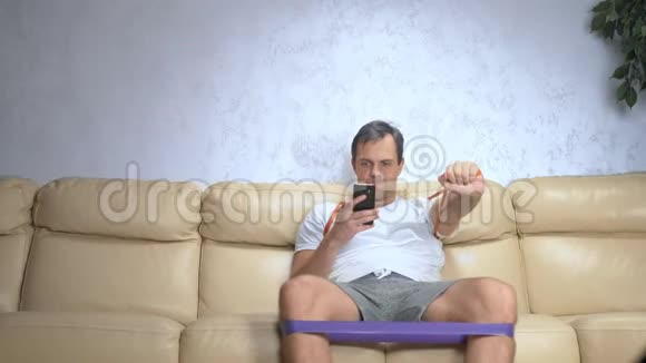 沙发上的男人用弹力带做运动用智能手机视频的预览图