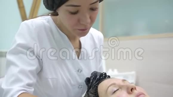 戴着刷子的粉红色手套的美容师用一个碳面膜在一个年轻女孩的脸上剥皮视频的预览图