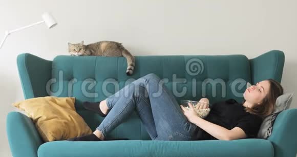女孩正在吃爆米花躺在家里的沙发上看电视侧视视频的预览图