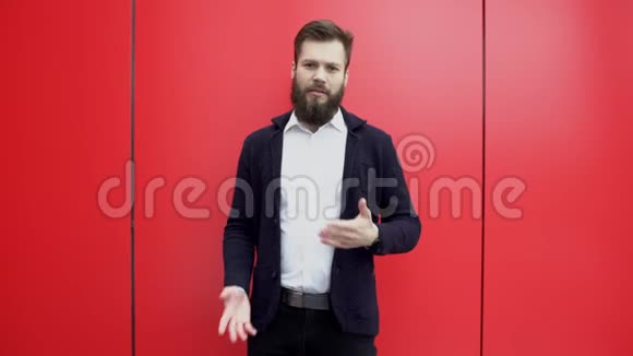 年轻的黑发胡子男子正在做手势并在红色背景下与相机交谈穿黑色夹克的视频的预览图