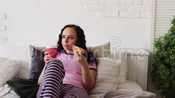 一个年轻的女人在床上吃热狗和喝咖啡在床上吃早餐把食物送到你家女人吃东西视频的预览图