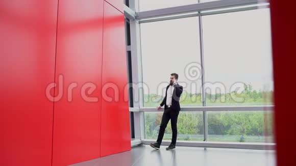 布吕内特留着胡子的商人正站在红墙房间的全景窗口旁打电话视频的预览图