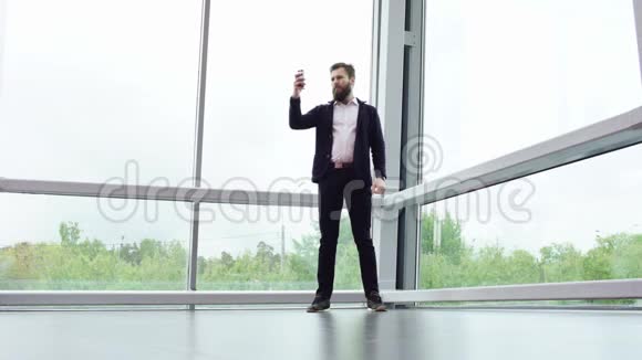 大胡子男子正在他的智能手机上拍照而站在现代建筑与大的全景窗口Brunette视频的预览图