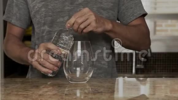 穿着灰色T恤的年轻人在一个漂亮时尚的厨房里用热水瓶或塑料瓶把水倒入玻璃杯中视频的预览图