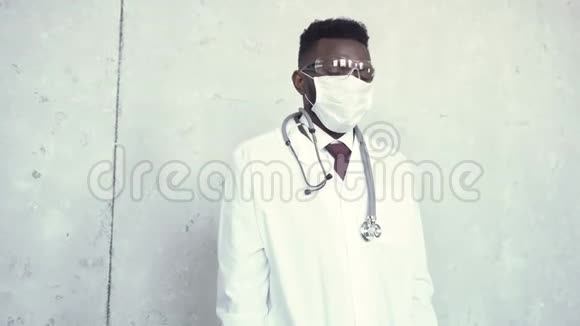 一位穿着白色实验室外套医用口罩和安全眼镜的黑人男性医生用蓝色手套擦拭他汗流浃背的前额视频的预览图