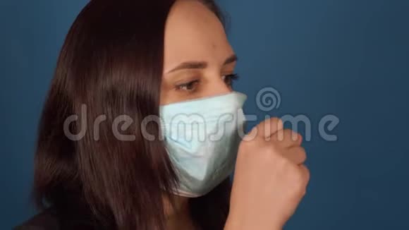 年轻女子戴着医用面具咳嗽的肖像在蓝色背景下被隔离在家里保护成年女性视频的预览图