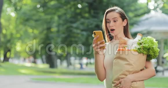 惊讶的女孩说哇和微笑同时检查新闻饲料和站在公园女人手里拿着一个装满食物的袋子视频的预览图