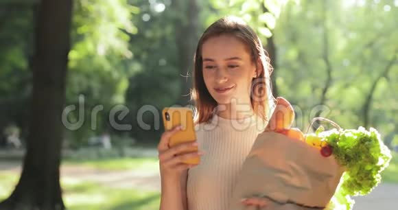 幸福的千禧一代女人使用她的智能手机拿着纸袋和食物漂亮开朗的女孩在看镜头视频的预览图