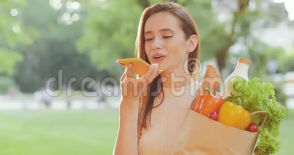 漂亮的年轻女人一边说话一边使用智能手机语音指挥官一边拿着带食物的纸袋快乐的千禧年视频的预览图