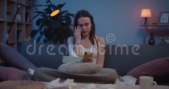 年轻沮丧的女人在拿着智能手机时使用纸组织的正面视图不开心的女孩哭着视频的预览图