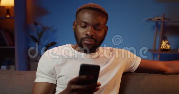 非洲帅哥坐在沙发上看智能手机屏幕的正面景色有胡子的年轻人在聊天视频的预览图