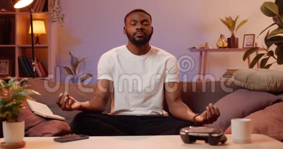 年轻的帅哥在家里坐在沙发上做呼吸锻炼在放松的时候做瑜伽的非洲男人视频的预览图