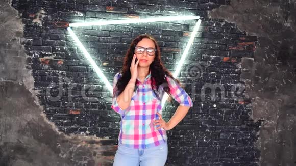 年轻女子戴着眼镜穿着彩色格子衬衫在背景破旧的砖墙上摆着发光的灯迷人的铁画像视频的预览图