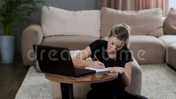 金发女孩在家里工作穿着一台漂亮的内部笔记本电脑在笔记本上写字视频的预览图