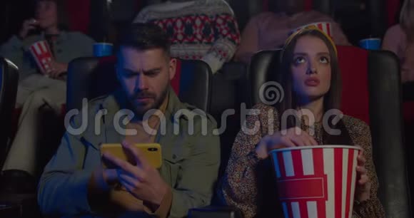 男人在电影院和女友约会时看体育比赛一个男人用智能手机一个女孩一边吃爆米花视频的预览图