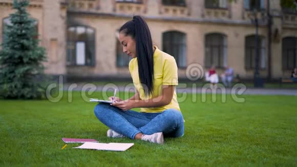 在校园里坐在草地上做作业的专注女学生在笔记本上做笔记4K视频的预览图