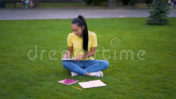 穿着黄色T恤的女学生坐在草地上正在为大学的考试做准备4K视频的预览图