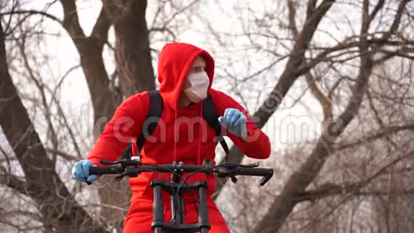 戴着口罩和手套的年轻人咳嗽坐在农村的自行车上穿红色西装保护成年男性视频的预览图