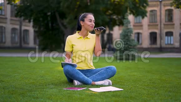 女学生坐在公园的草地上倾听并记录一条语音信息4K视频的预览图