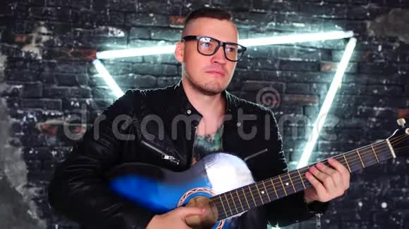 纹身男在照明墙附近弹吉他时髦的男人胸前有鸟纹看着远处弹吉他视频的预览图