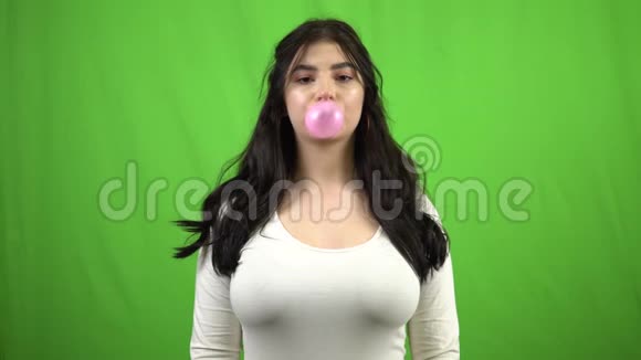 年轻的黑发女孩在绿色屏幕上吹泡泡糖看着相机视频的预览图