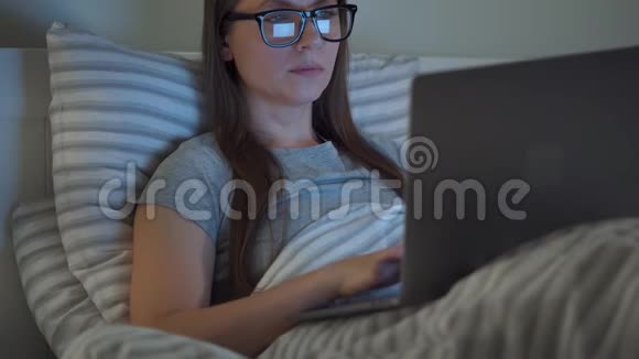 戴眼镜的女人晚上躺在床上用笔记本电脑工作网络成瘾或失眠的概念视频的预览图