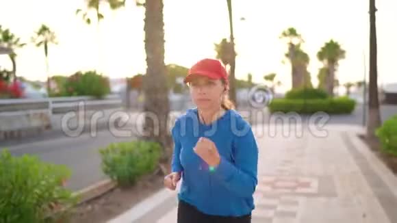 在日落时分戴着耳机的女人沿着棕榈大道沿着街道奔跑健康的生活方式视频的预览图