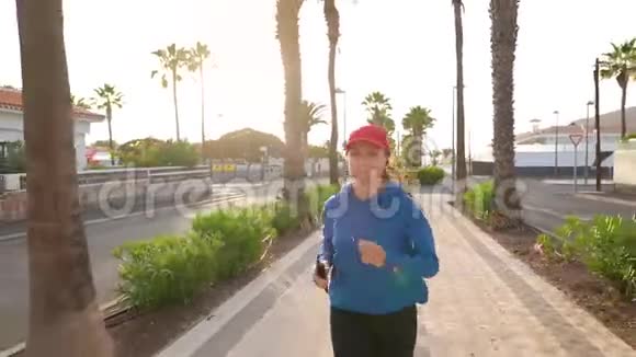 日落时分带着耳机和智能手机的女人沿着棕榈大道沿着街道跑健康的生活方式视频的预览图
