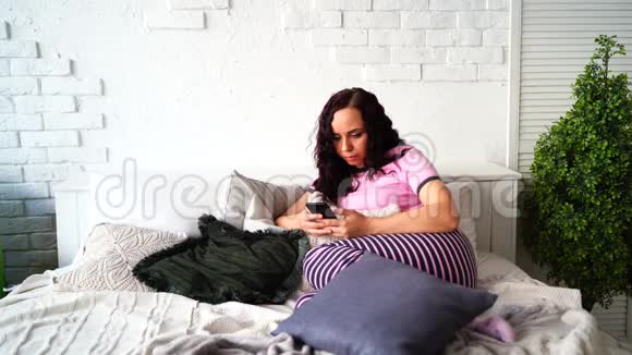 穿着睡衣的成年迷人女性在卧室的手机里翻着各种新闻年轻女子远程学习视频的预览图