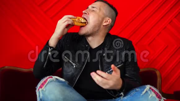 英俊的年轻人吃汉堡躺在红色墙上的小沙发上成年男性脚坐着享受食物视频的预览图