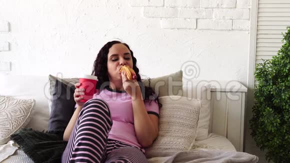 一个年轻的女人在床上吃热狗和喝咖啡在床上吃早餐把食物送到你家女人吃东西视频的预览图