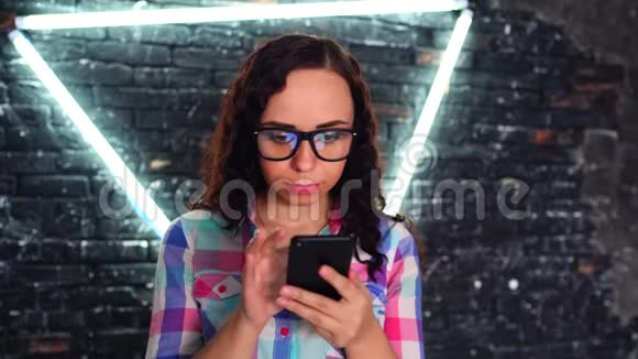 戴着眼镜的年轻美女手拿着手机站在背景砖墙上放着发光的灯视频的预览图