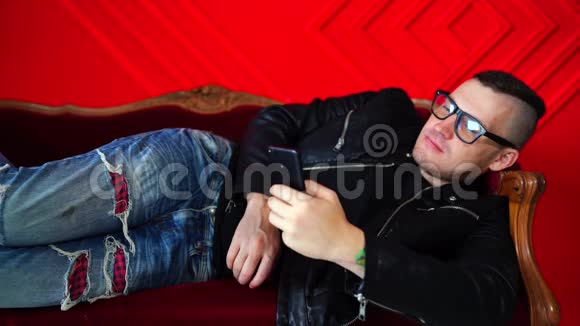 英俊的年轻人戴着眼镜坐在手机里靠在红墙的小沙发上成年迷人男性翻转视频的预览图
