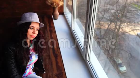 戴着帽子和皮夹克的年轻美女坐在窗户旁从窗户向外看的漂亮女人的肖像视频的预览图