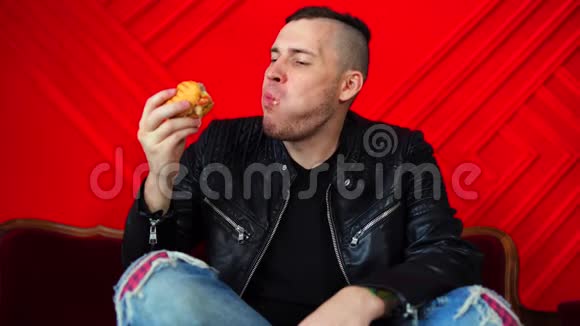 英俊的年轻人吃汉堡躺在红色墙上的小沙发上成年男性脚坐着享受食物视频的预览图