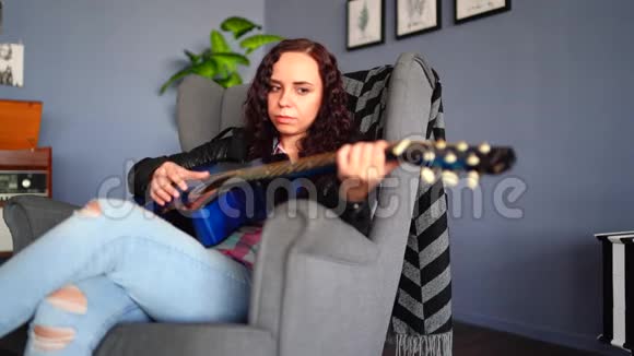 年轻漂亮的女人穿着便服坐在扶手椅上在家里弹吉他漂亮女性的肖像视频的预览图