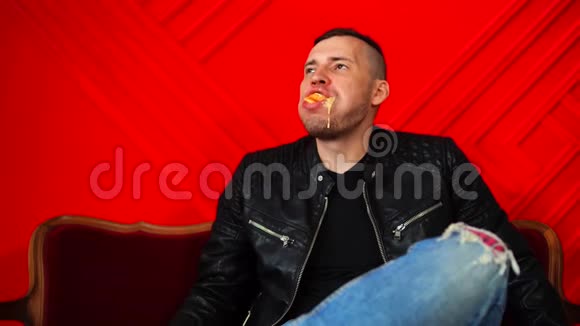 英俊的年轻人咀嚼食物满嘴都是靠在红墙的小沙发上成年男性享受食物视频的预览图