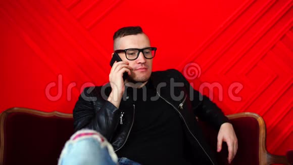 英俊的年轻人戴着眼镜拿着智能手机躺在红色墙上的小沙发上成年迷人男性在交谈视频的预览图