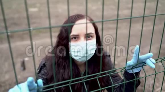 年轻女子戴着医用口罩通过栅栏遮住脸成年女性用面具遮住她的脸视频的预览图