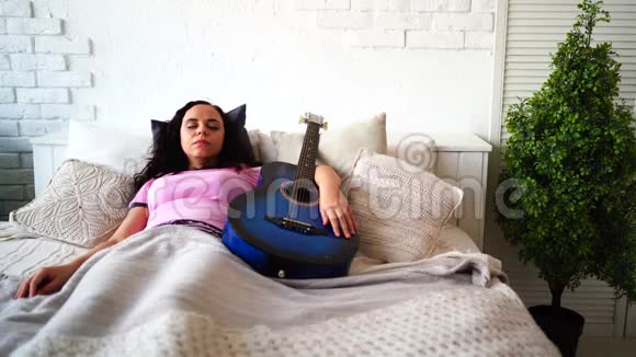 年轻女子抱着吉他在房间里休息穿着睡衣睡着乐器的成年女性视频的预览图