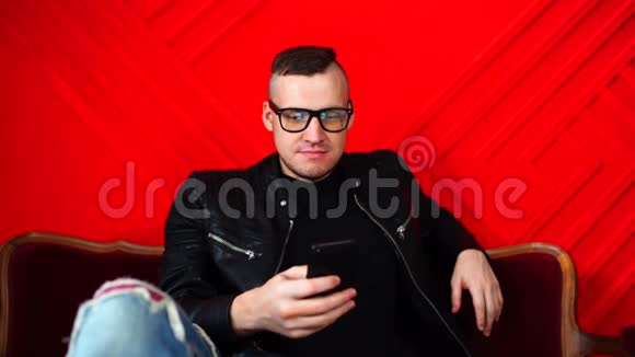 英俊的年轻人戴着眼镜拿着智能手机躺在红色墙上的小沙发上成年男性坐扶手椅视频的预览图