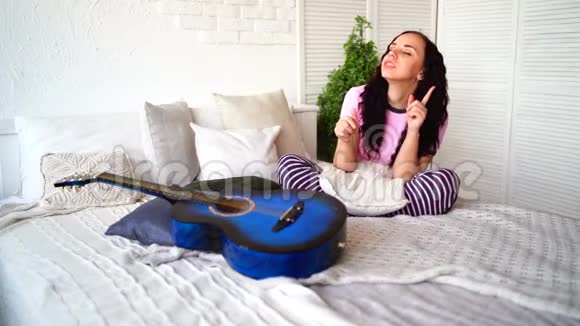 年轻的女人坐在房间里拿着吉他享受她的放松成年女性穿着睡衣跳舞坐在床上视频的预览图