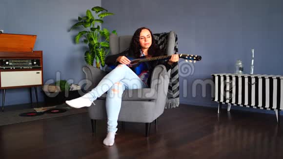 年轻漂亮的女人穿着便服坐在扶手椅上在家里弹吉他漂亮女性的肖像视频的预览图