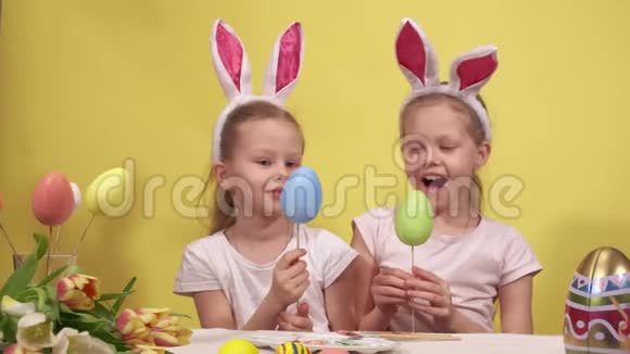 快乐的小女孩带着小兔子的耳朵微笑着在木棍上演示彩蛋坐在桌子上准备视频的预览图