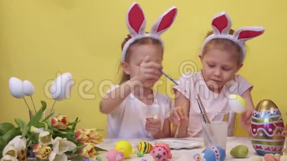 专注的姐妹兔子耳朵站在桌子附近调色板和郁金香花束仔细着色鸡蛋而视频的预览图
