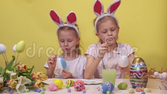 专注的姐妹兔子耳朵站在桌子附近调色板和郁金香花束仔细着色鸡蛋而视频的预览图