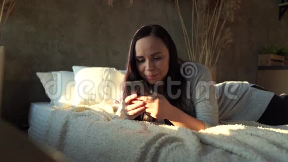 穿着休闲服躺在床上的漂亮年轻女人成人微笑的女士在手机里翻阅各种新闻视频的预览图