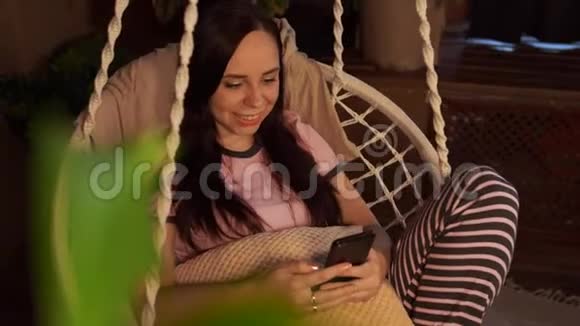 快乐的年轻女人躺在吊床上坐在家里的智能手机上年轻迷人的年轻女性看相机视频的预览图