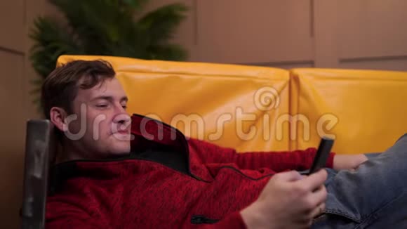 穿休闲服装的年轻人躺在黄色沙发上成年男性在手机中休息和翻阅各种新闻视频的预览图